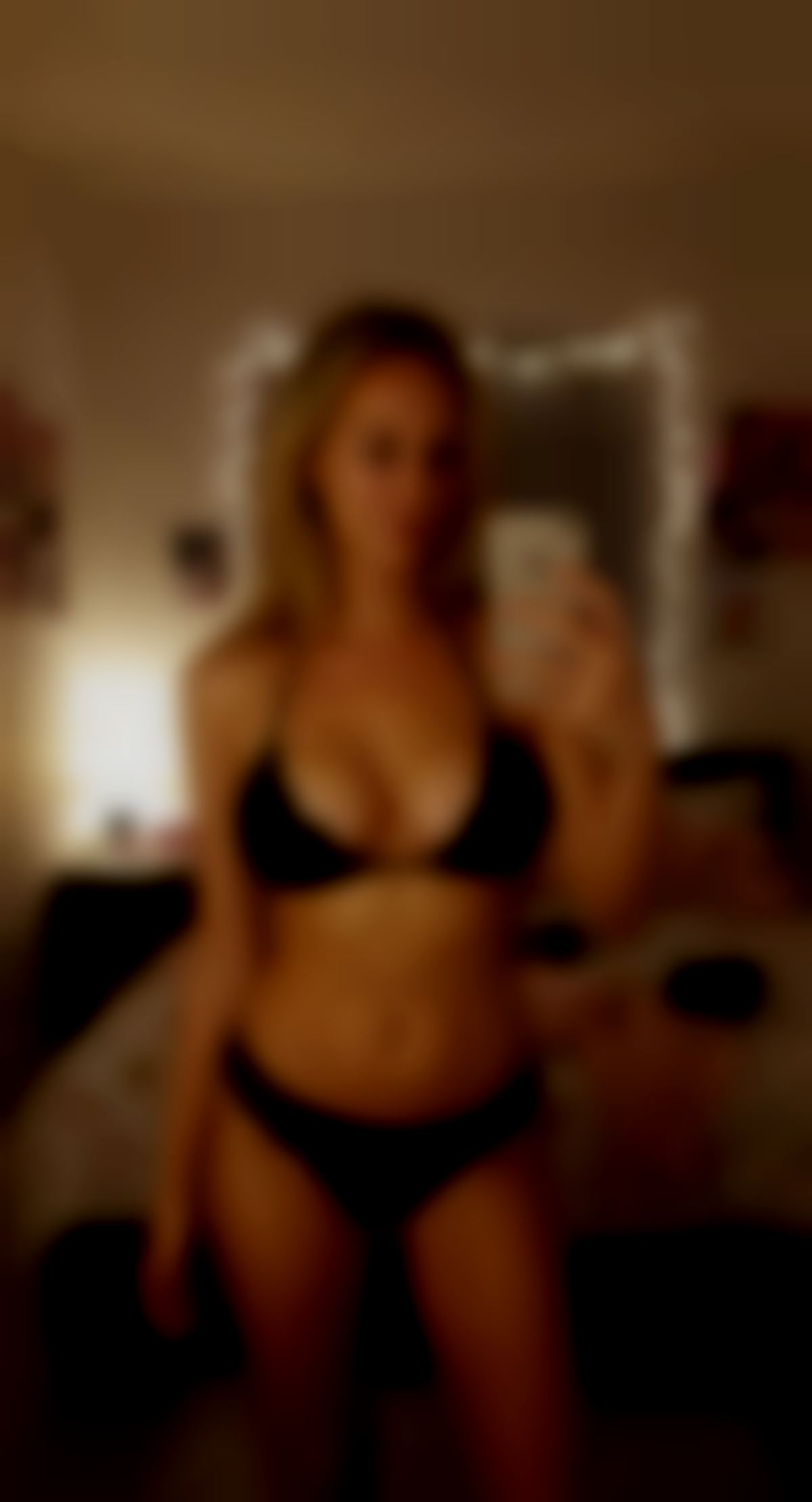 A beautiful blonde woman standing in her bedroom, holding her phone in her right hand.