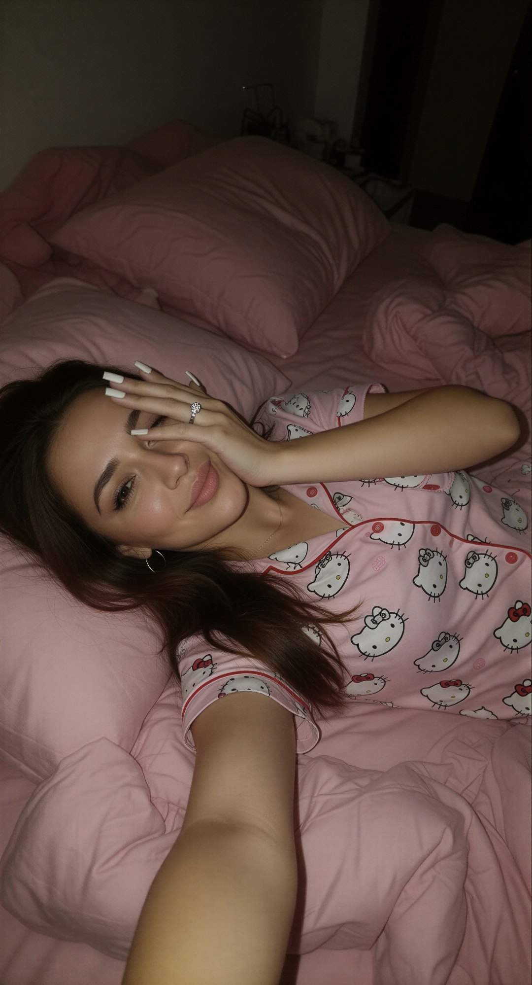 Edit the reference image: lying sideways on her left side on a messy pink bed in a dimly lit bedroom at night, wearing soft pink Hello Kitt…