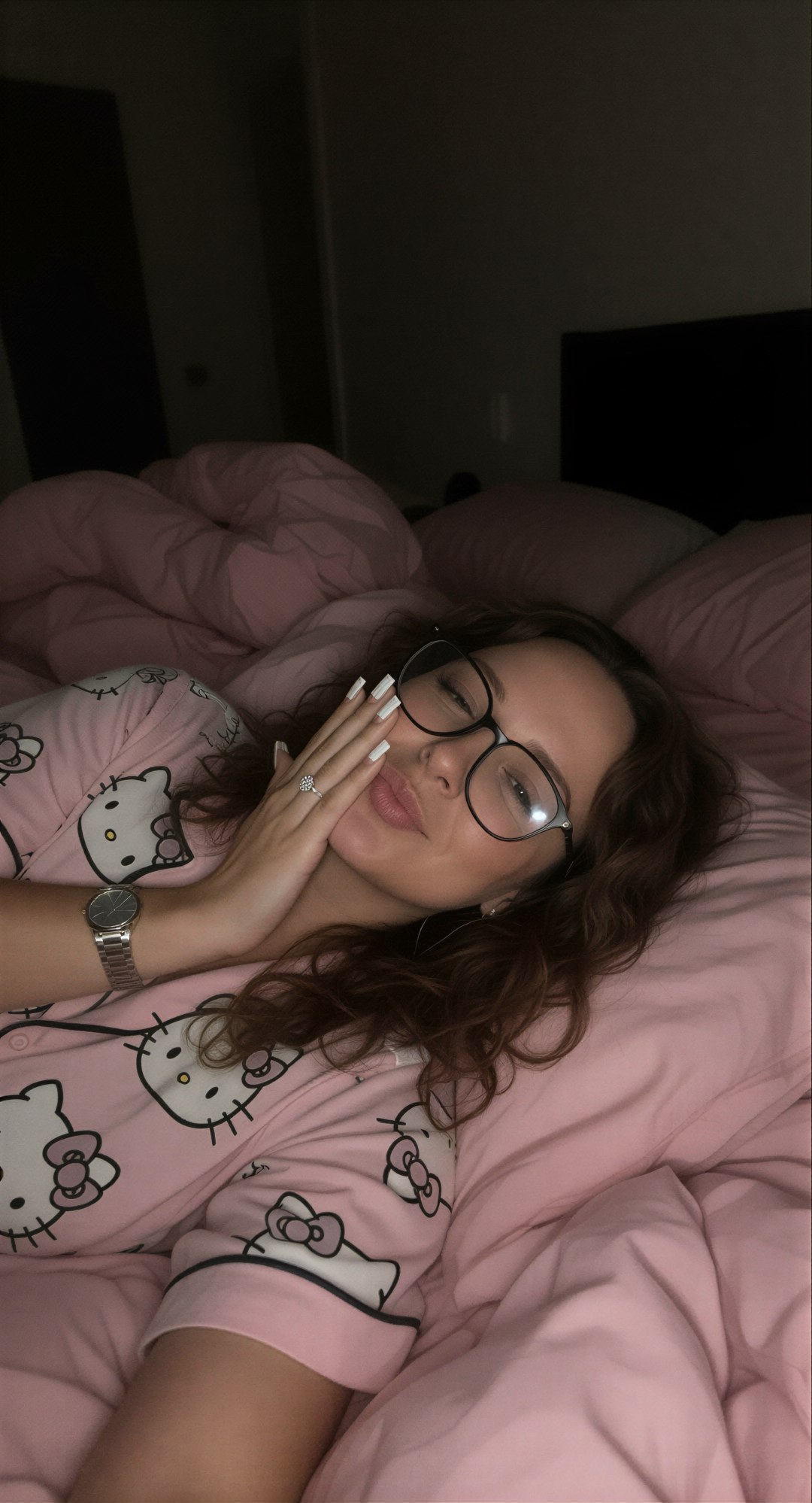 Edit the reference image: lying sideways on her left side on a messy pink bed in a dimly lit bedroom at night, wearing soft pink Hello Kitt…