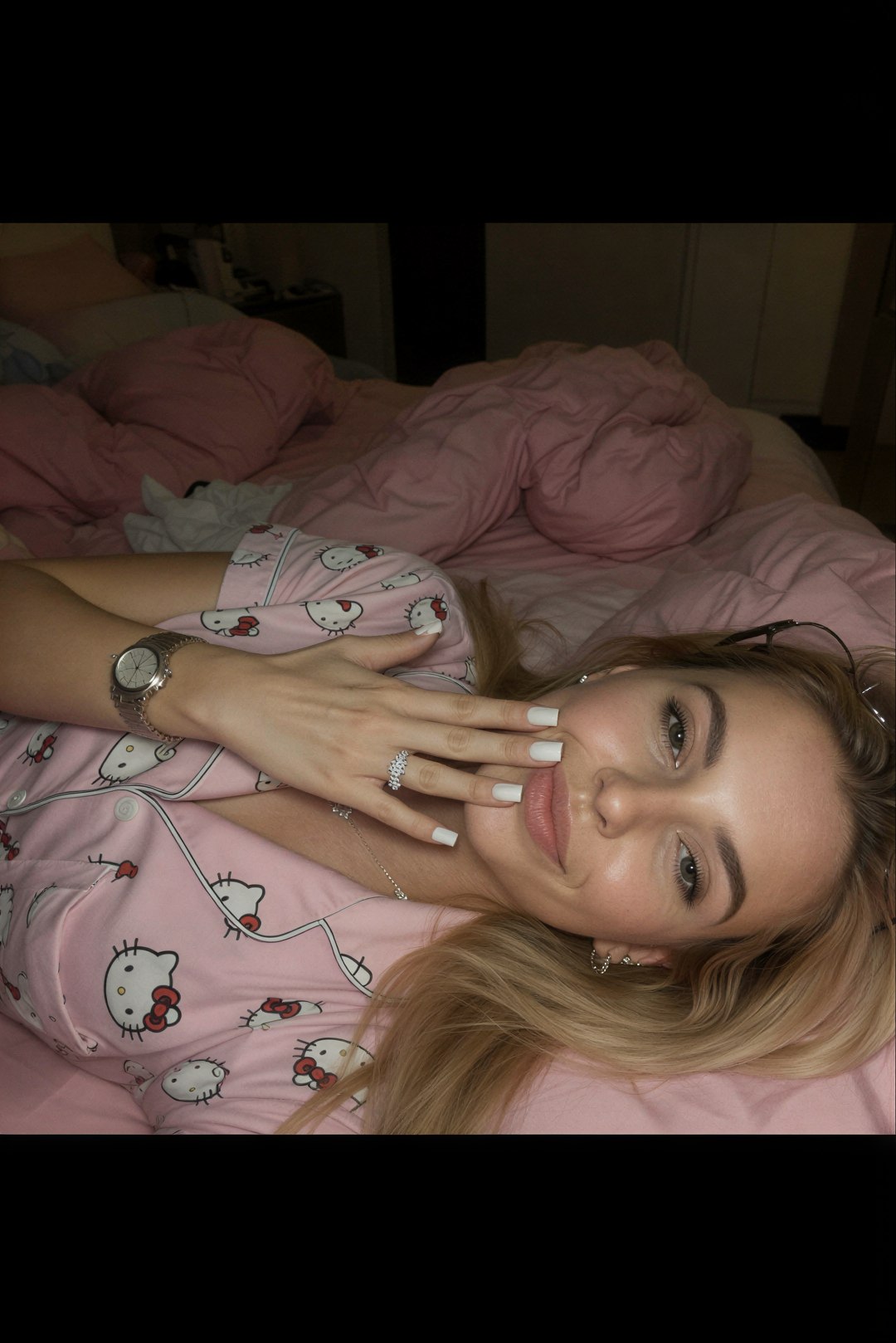 Edit the reference image: lying sideways on her left side on a messy pink bed in a dimly lit bedroom at night, wearing soft pink Hello Kitt…